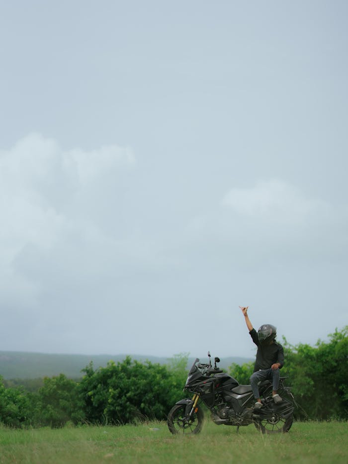 A motorcyclist raises hand triumphantly on a bike in a scenic outdoor landscape.
