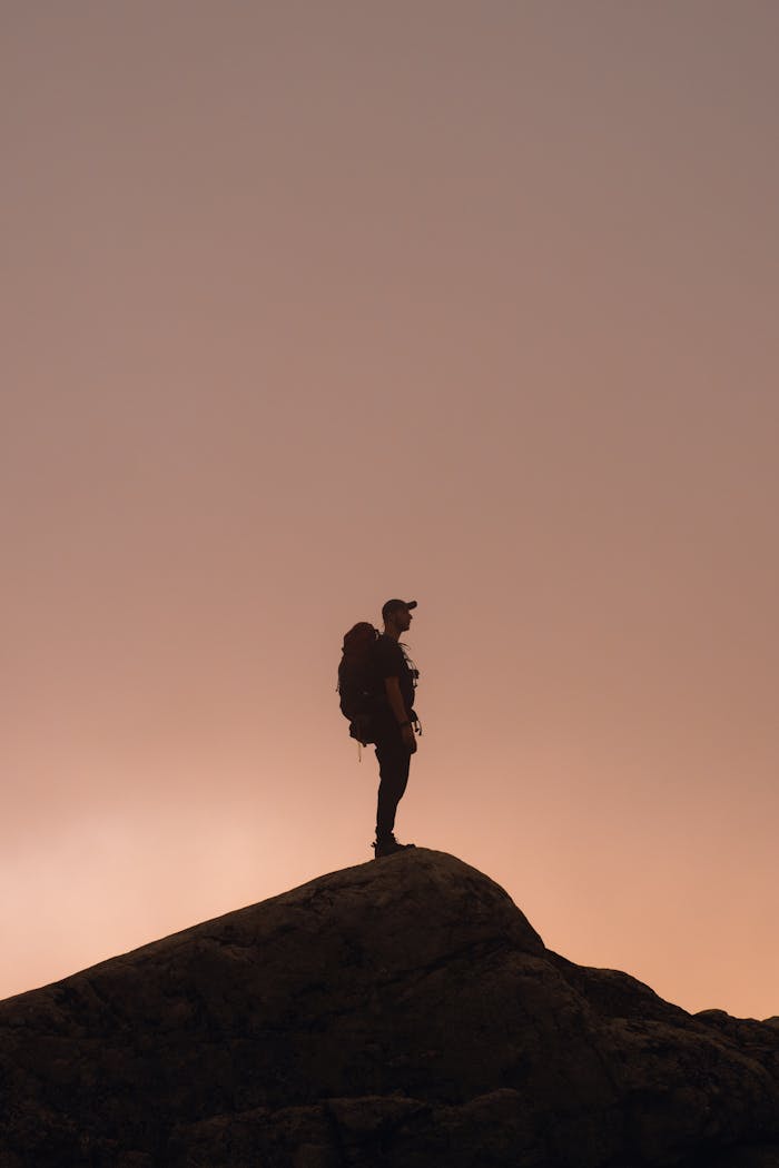 our-stories A lone hiker silhouetted against a dramatic sunset sky on a mountain peak, symbolizing adventure.