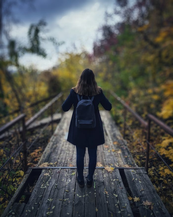 A young woman with a backpack stands on a wooden bridge surrounded by vibrant fall foliage.